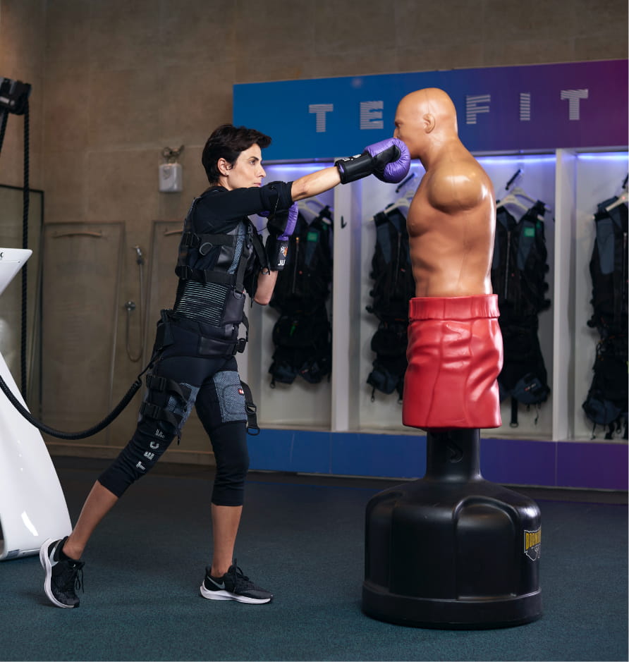 Aluna com traje de eletroestimulação golpeando um boneco de boxe dentro do estúdio Tecfit